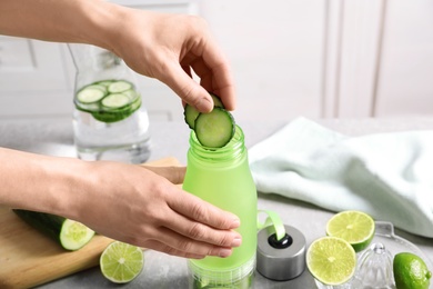 Young woman putting fresh cucumber slices into sports bottle on table. Space for text