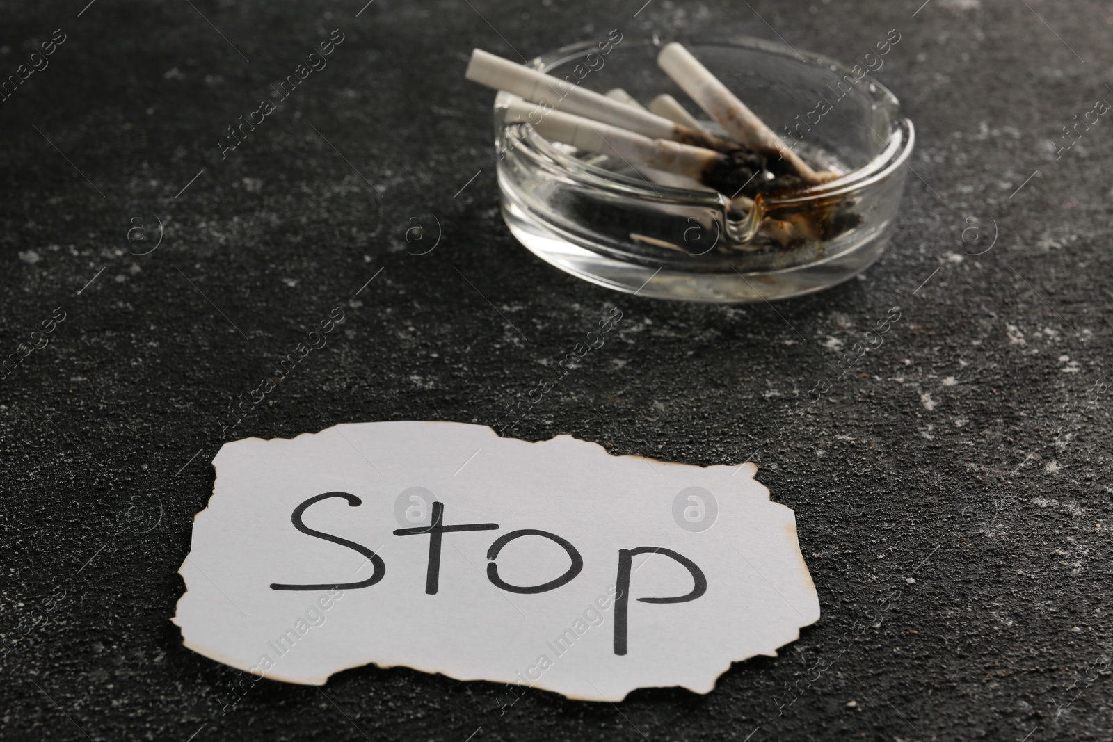 Photo of Ashtray with burnt cigarettes and word Stop written on paper on black textured table, closeup. No smoking concept