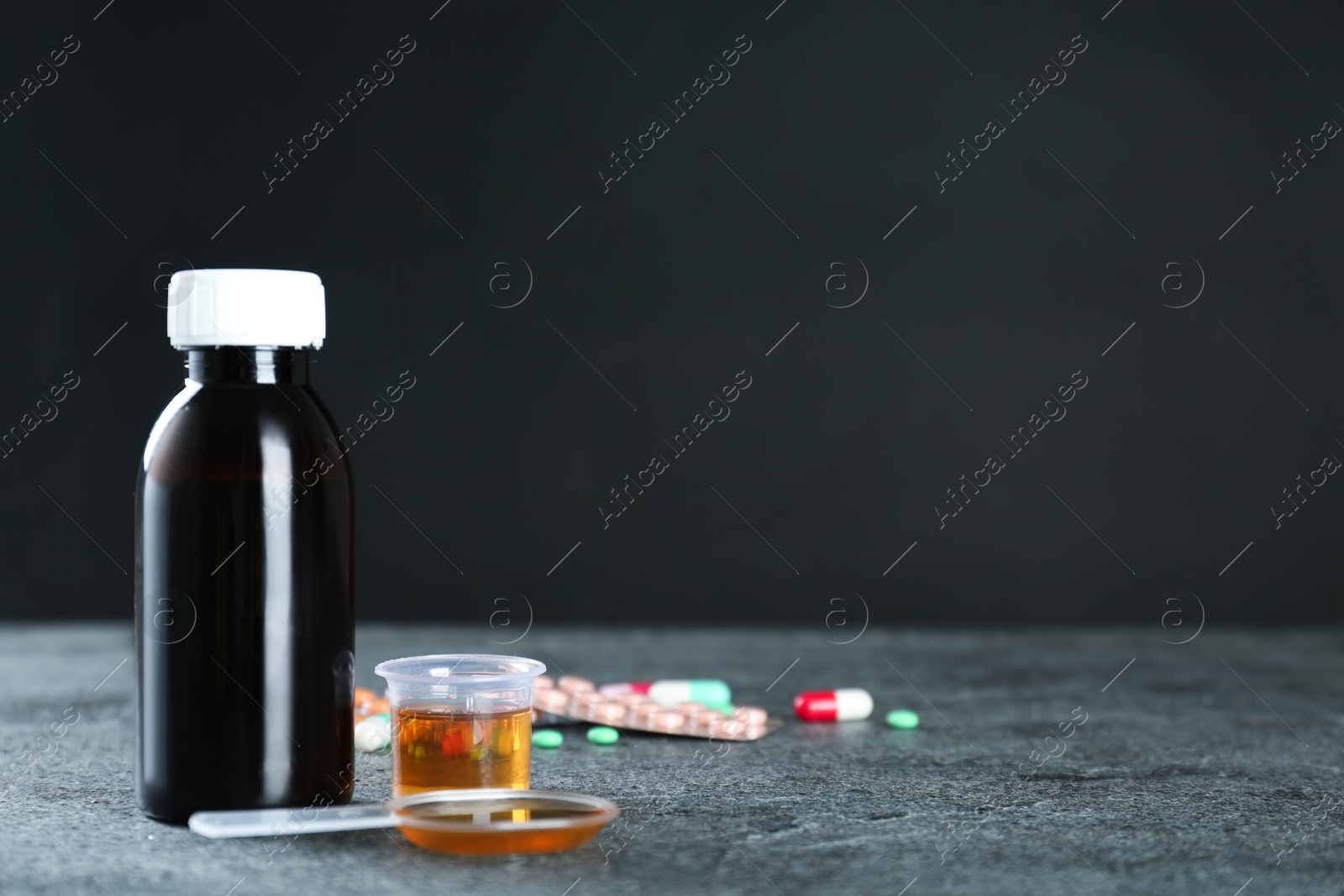 Photo of Bottle of cough syrup, dosing spoon and measuring cup on grey table. Space for text