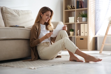 Happy woman reading letter while sitting on floor near sofa at home