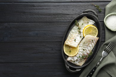 Photo of Fresh raw cod fillets with thyme and lemon in baking dish on black wooden table, flat lay. Space for text