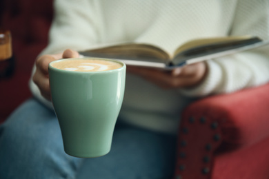 Woman with coffee reading book indoors, focus on cup