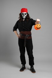 Man in scary pirate costume with skull makeup and pumpkin bucket on light grey background. Halloween celebration