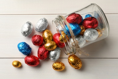 Overturned glass jar with chocolate eggs wrapped in colorful foil on white wooden table, flat lay