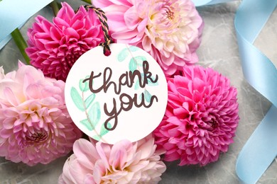 Photo of Paper tag with phrase Thank You and dahlias on marble table, flat lay