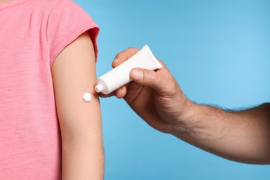 Father applying ointment onto his daughter's arm on light blue background, closeup