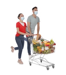 Couple with protective masks and shopping cart full of groceries on white background