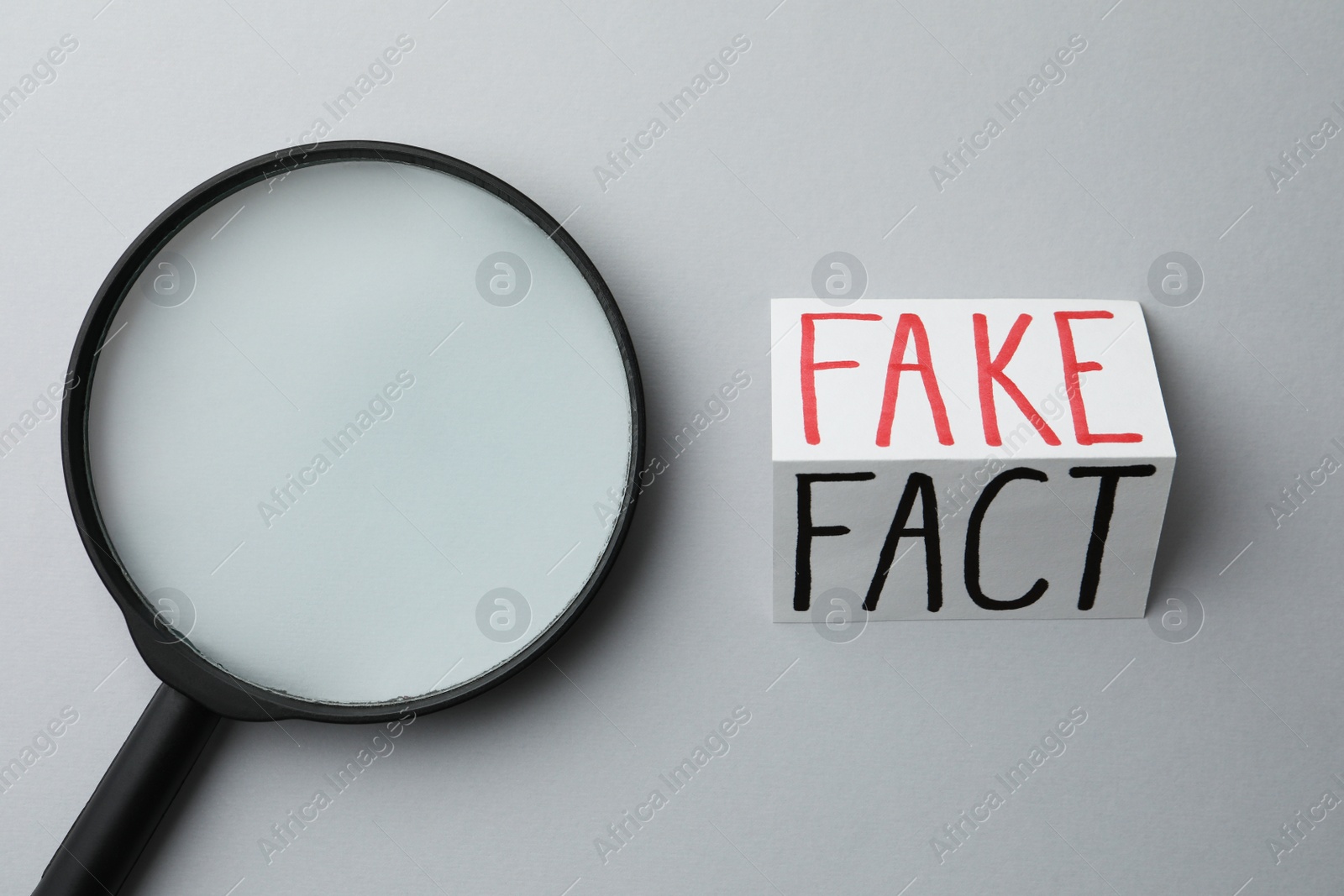 Photo of Magnifying glass near sheet of paper with words Fact and Fake on light grey background, flat lay