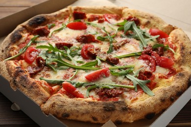 Photo of Tasty pizza with cheese, tomato and arugula in cardboard box on table, closeup