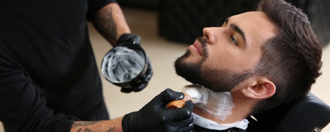 Image of Professional hairdresser applying shaving foam onto client's skin in barbershop. Banner design