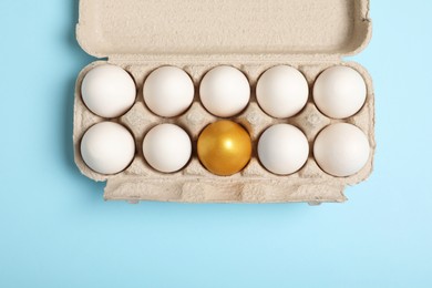 Cardboard box with golden and ordinary chicken eggs on light blue background, top view