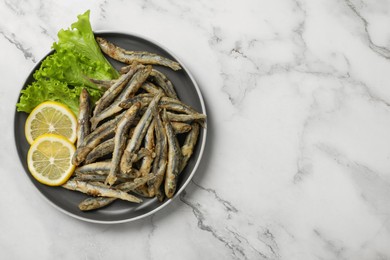 Photo of Plate with delicious fried anchovies, lemon and lettuce leaves on white marble table, top view. Space for text