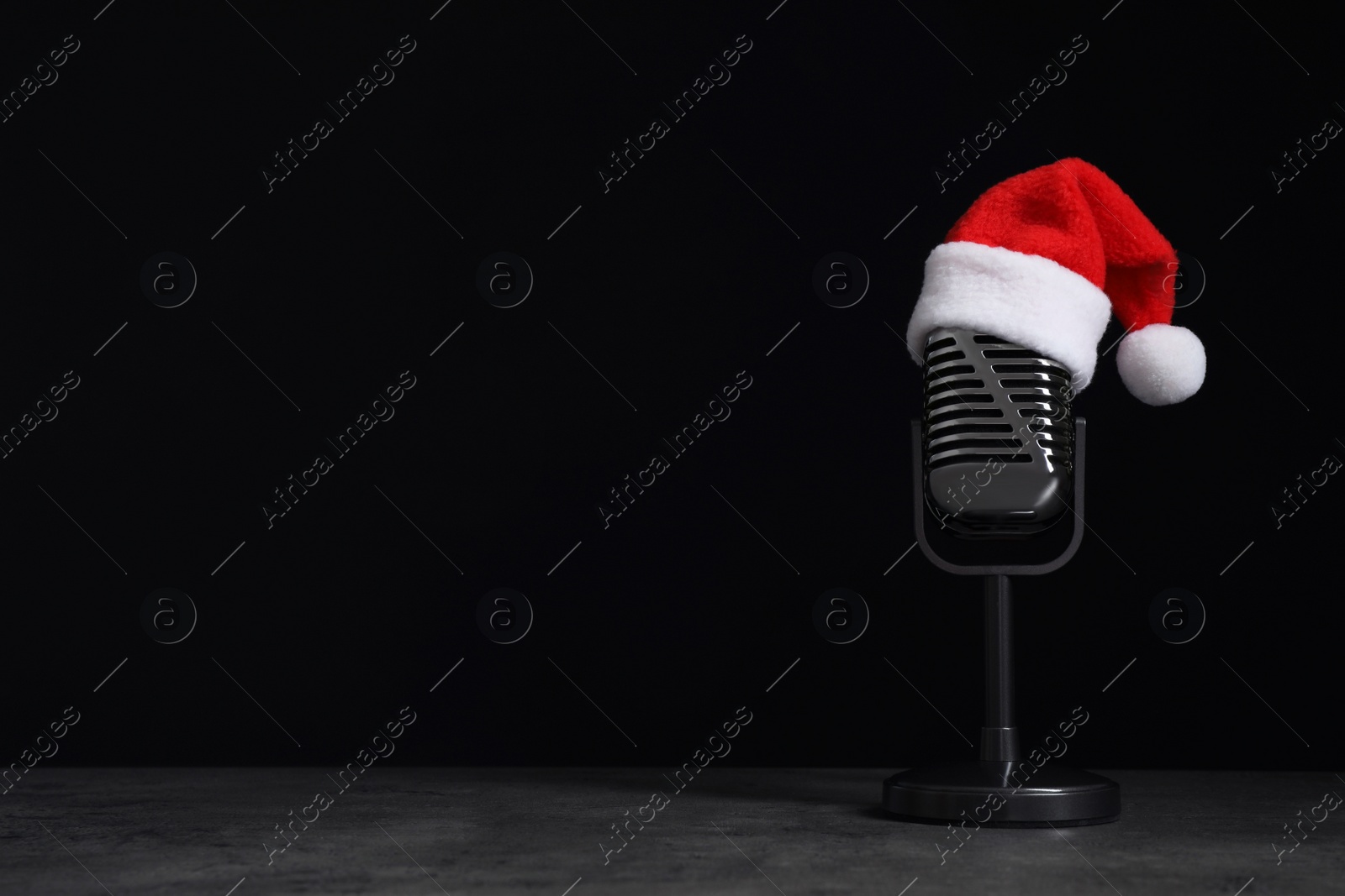 Photo of Microphone with Santa hat on grey stone table against black background, space for text. Christmas music