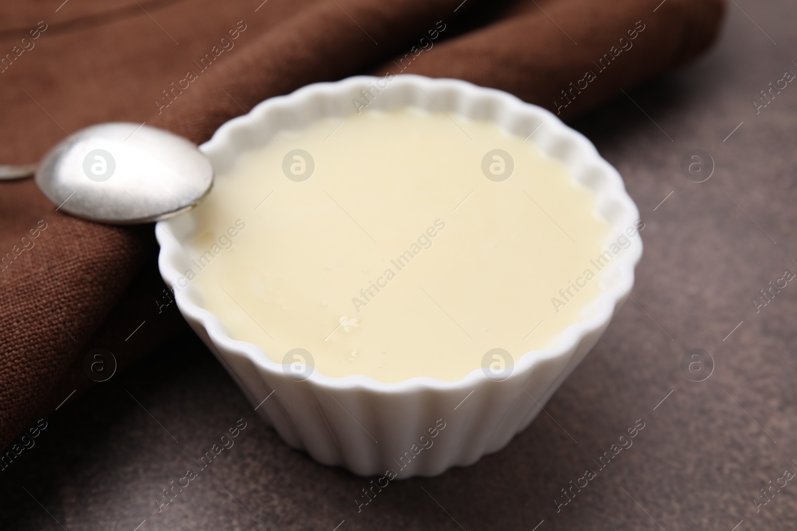 Photo of Bowl with condensed milk and spoon on brown table, closeup