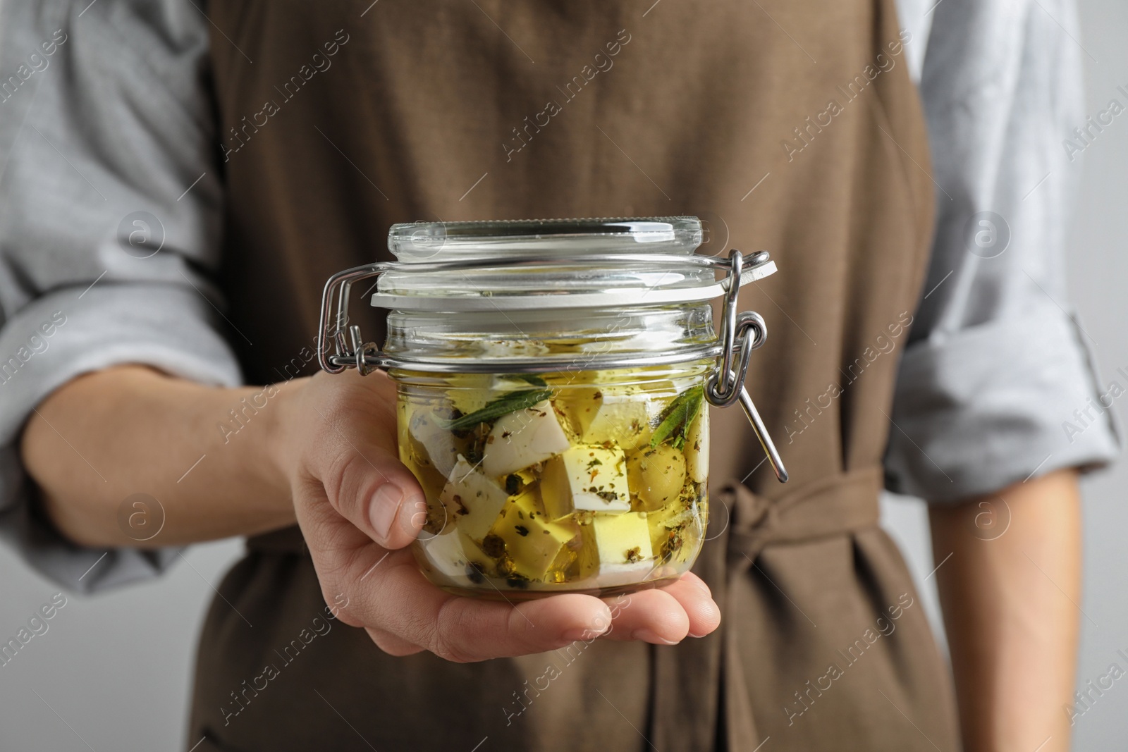 Photo of Woman holding jar with pickled feta cheese on grey background, closeup