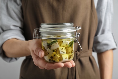 Woman holding jar with pickled feta cheese on grey background, closeup