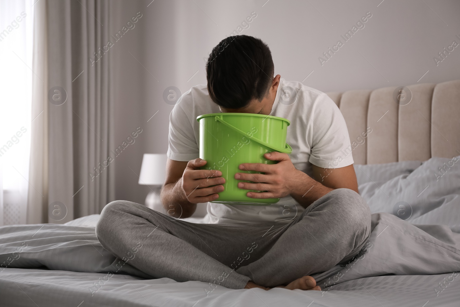 Photo of Man with bucket suffering from nausea on bed at home. Food poisoning