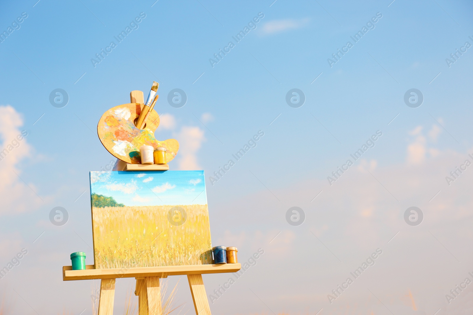 Photo of Wooden easel with beautiful picture and painting equipment against blue sky. Space for text