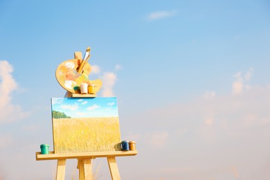 Wooden easel with beautiful picture and painting equipment against blue sky. Space for text