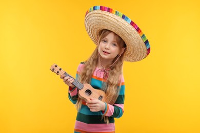 Photo of Cute girl in Mexican sombrero hat playing ukulele on orange background