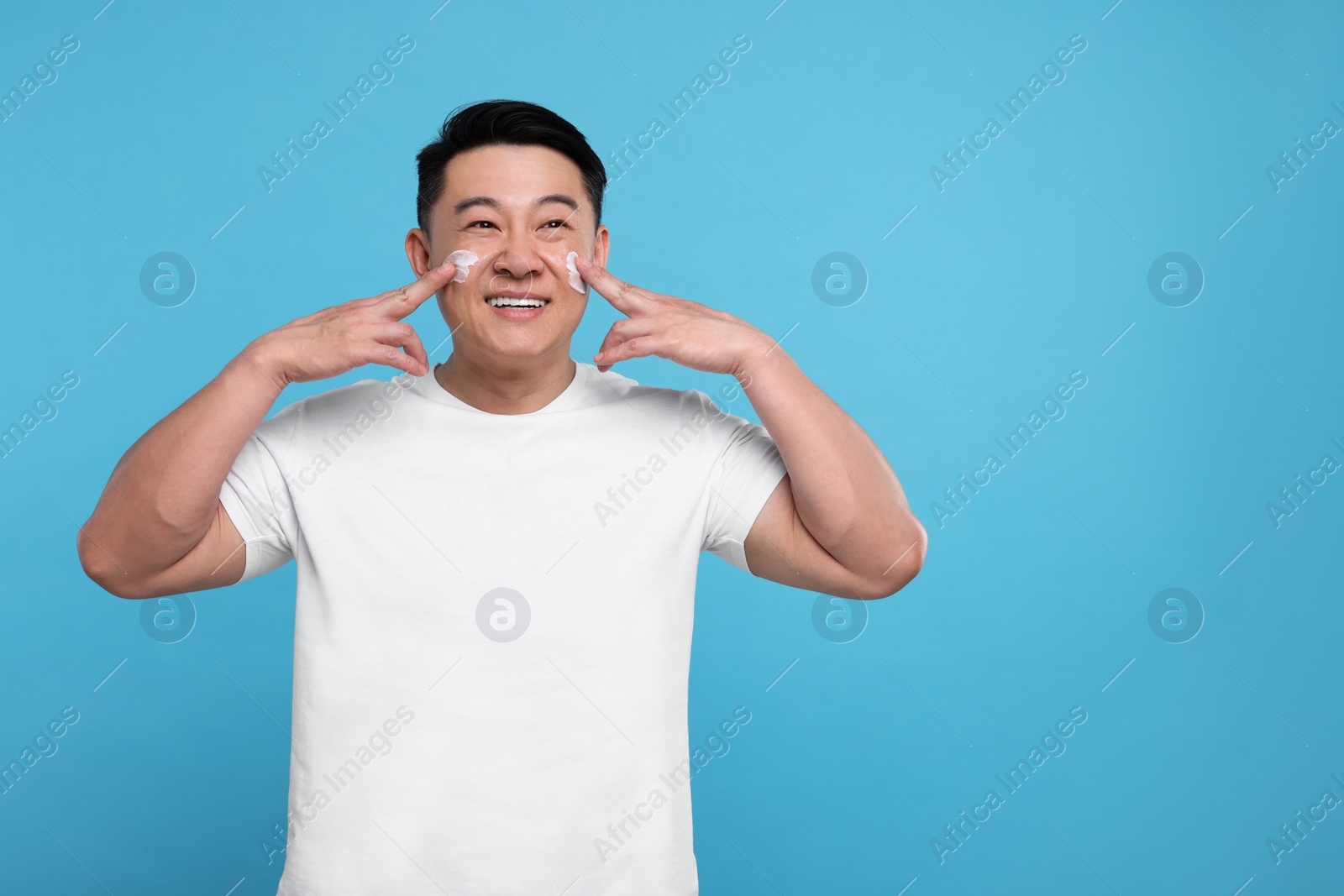 Photo of Handsome man applying cream onto his face on light blue background. Space for text