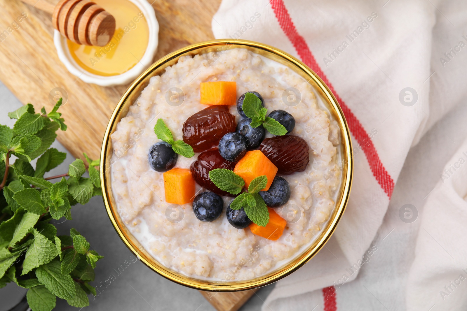Photo of Delicious barley porridge with blueberries, pumpkin, dates and mint served with honey on light table, flat lay