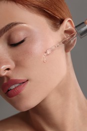 Photo of Beautiful young woman applying cosmetic serum onto her face on grey background, closeup