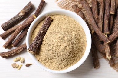 Powder in bowl and dried sticks of liquorice root on white table, flat lay