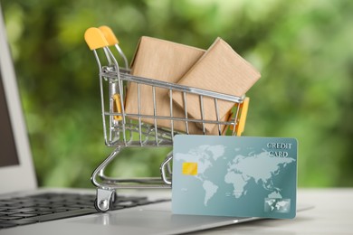 Online payment concept. Small shopping cart with bank card, boxes and laptop on table, closeup