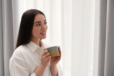 Happy woman with cup of drink near window at home, space for text. Lazy morning