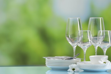 Photo of Set of many clean dishware, cutlery, flowers and glasses on light blue table against blurred green background. Space for text