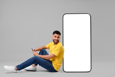 Man with mobile phone sitting near huge device with empty screen on grey background. Mockup for design