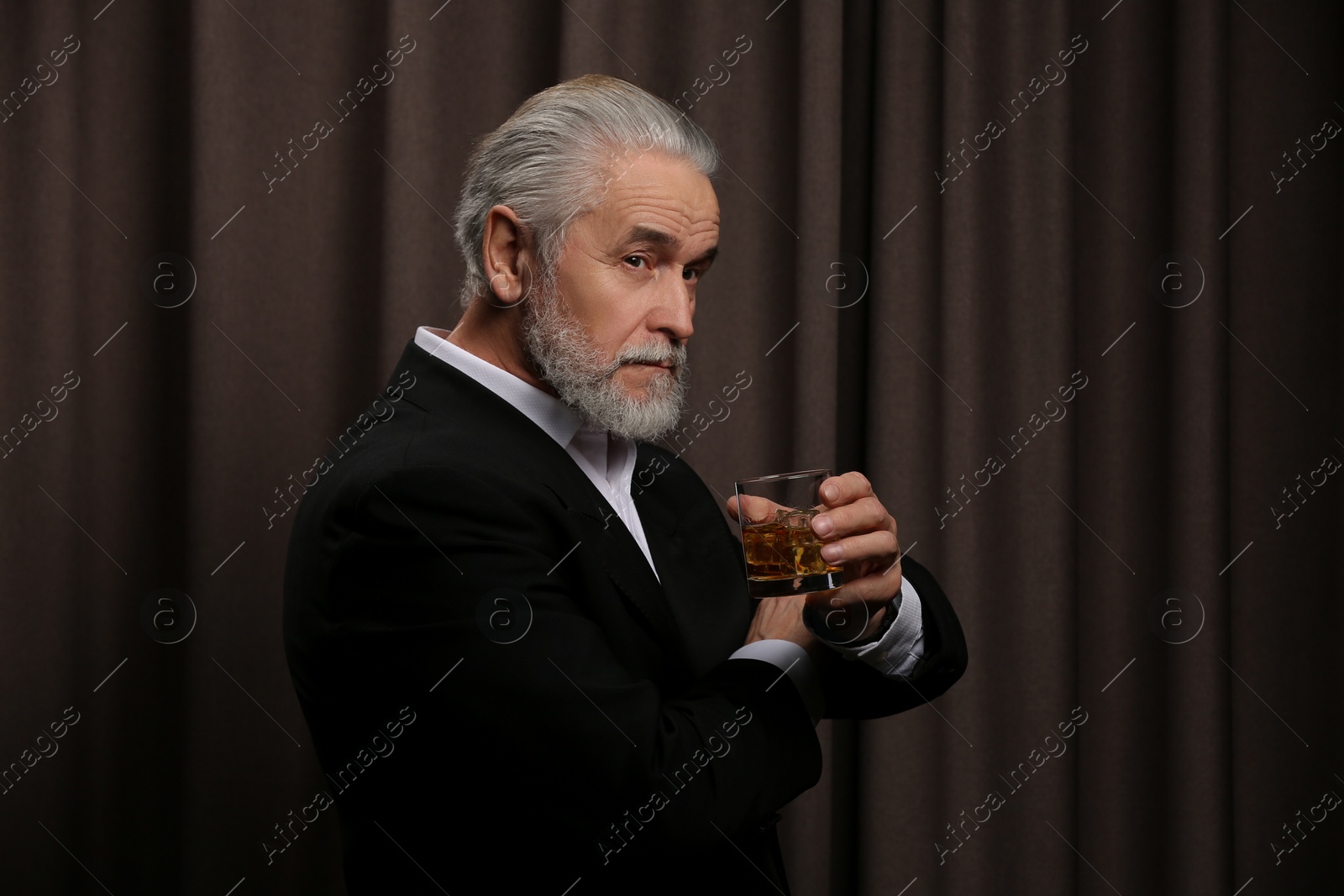 Photo of Senior man in suit holding glass of whiskey with ice cubes on brown background. Space for text