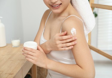 Young woman applying body cream on shoulder indoors, closeup