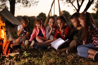 Photo of Little children reading book with flashlight outdoors. Summer camp