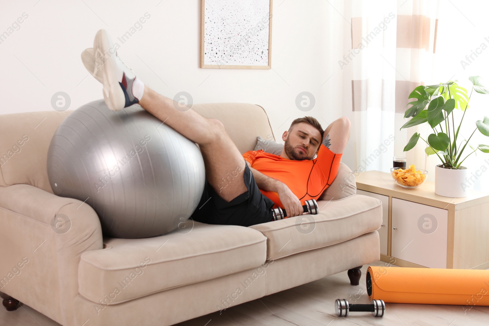 Photo of Lazy young man with sport equipment on sofa at home