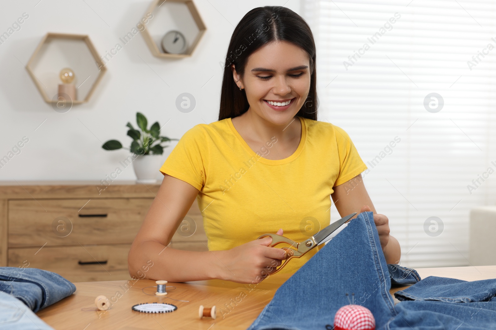 Photo of Young woman cutting jeans with scissors at wooden table indoors