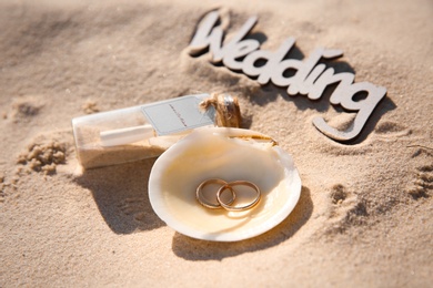 Photo of Shell with gold rings, invitation in glass bottle and word Wedding on sandy beach, closeup