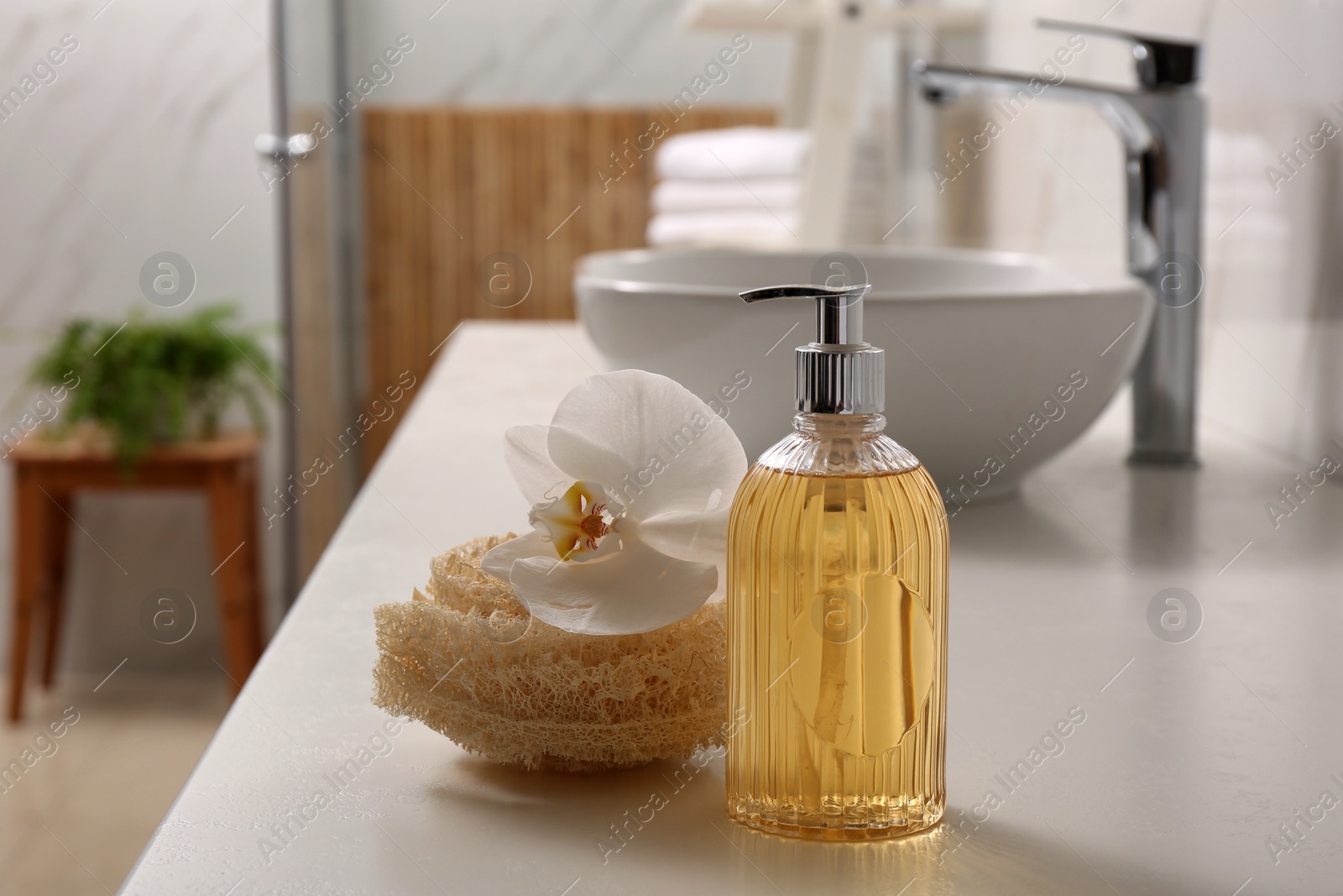 Photo of Glass dispenser with liquid soap, loofah and beautiful flower on white table in bathroom