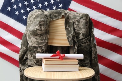 Diploma, books and soldier uniform on wooden chair near flag of United States. Military education