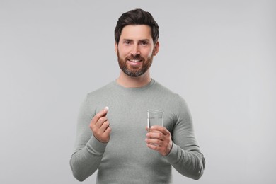 Handsome man with glass of water and pill on light grey background