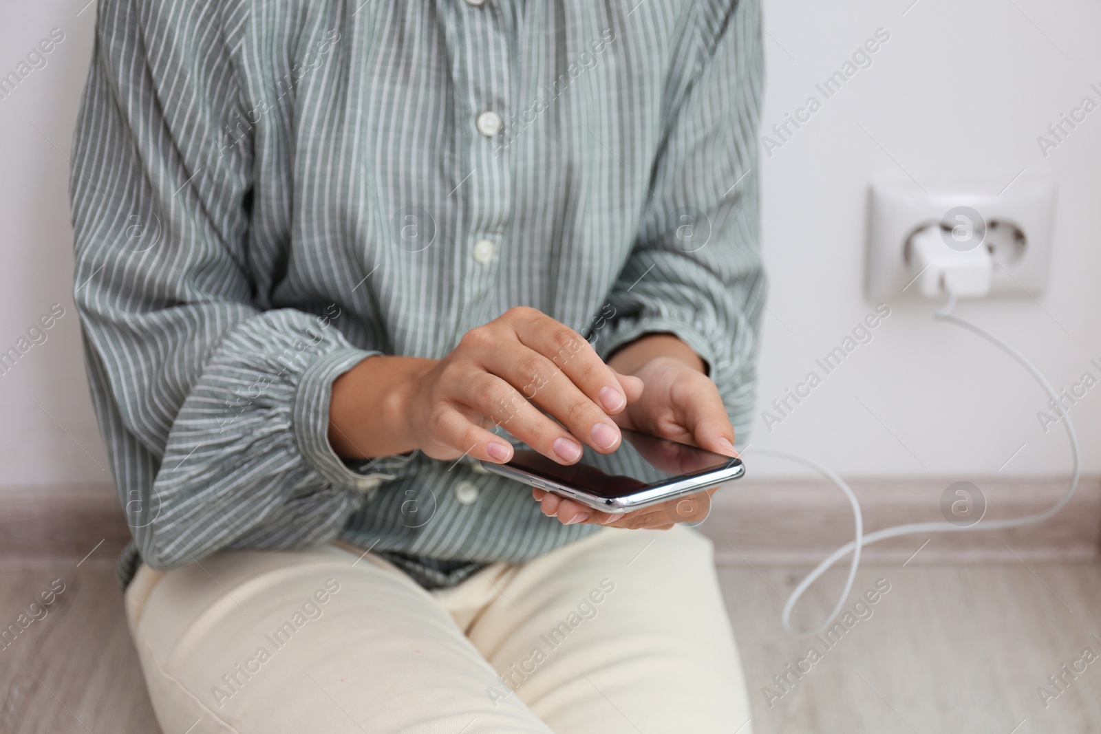 Photo of Woman using mobile phone with connected charger near white brick wall indoors, closeup