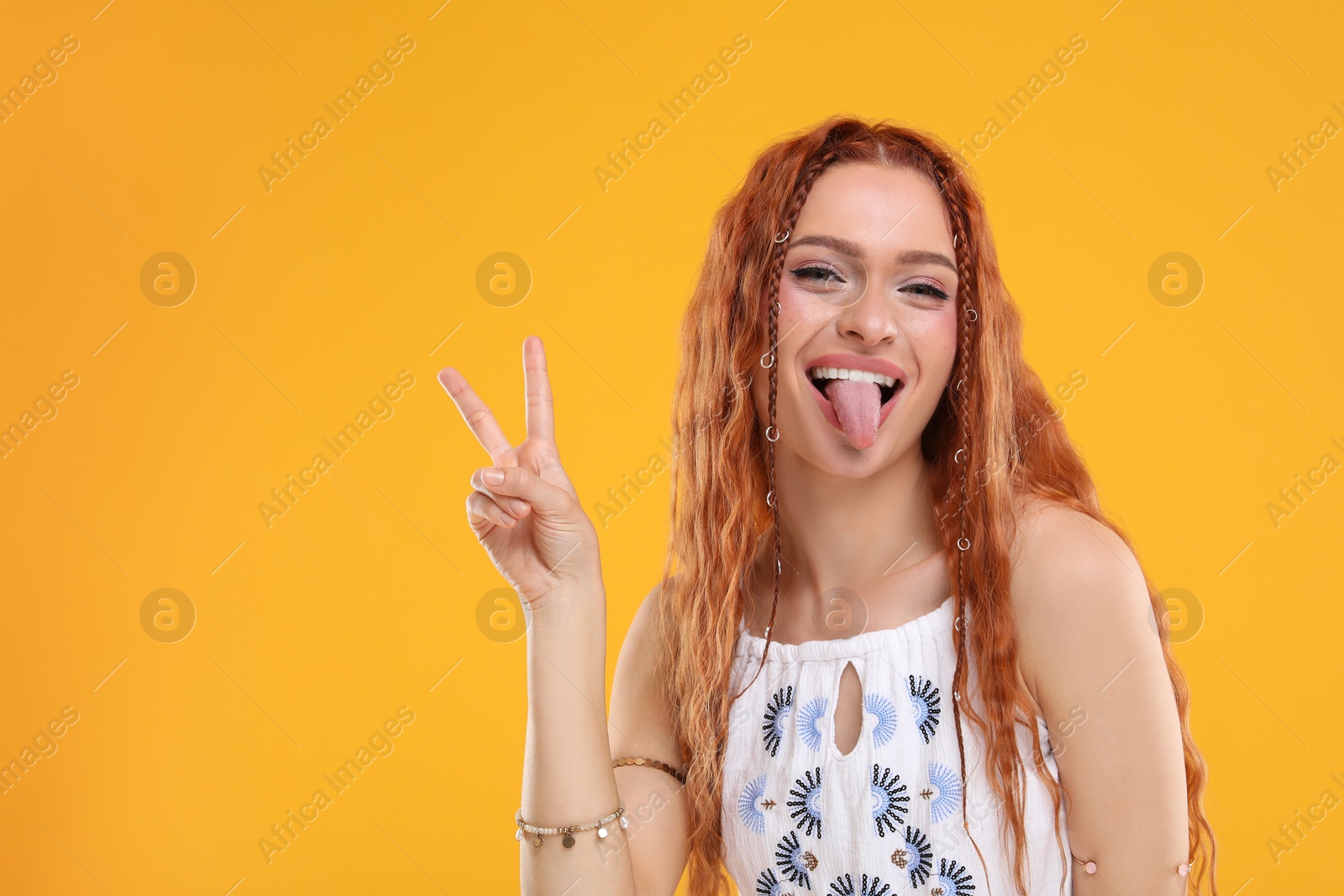 Photo of Beautiful young hippie woman showing her tongue and V-sign on orange background, space for text