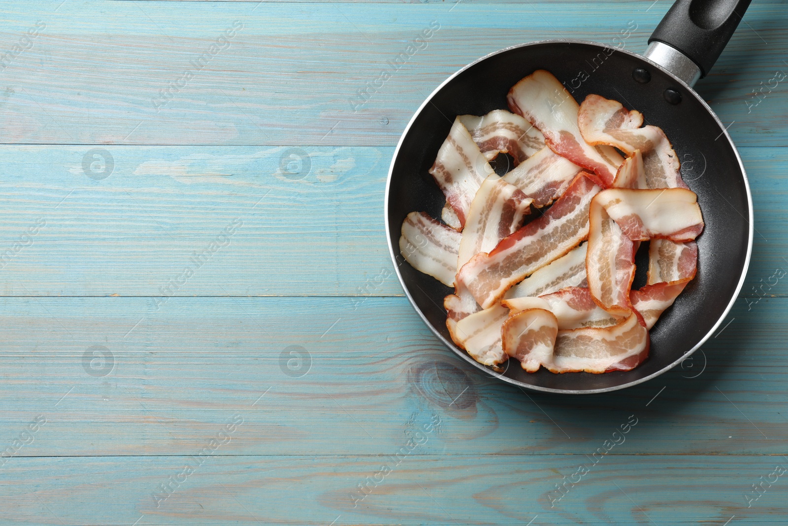 Photo of Delicious bacon slices in frying pan on light blue wooden table, top view. Space for text