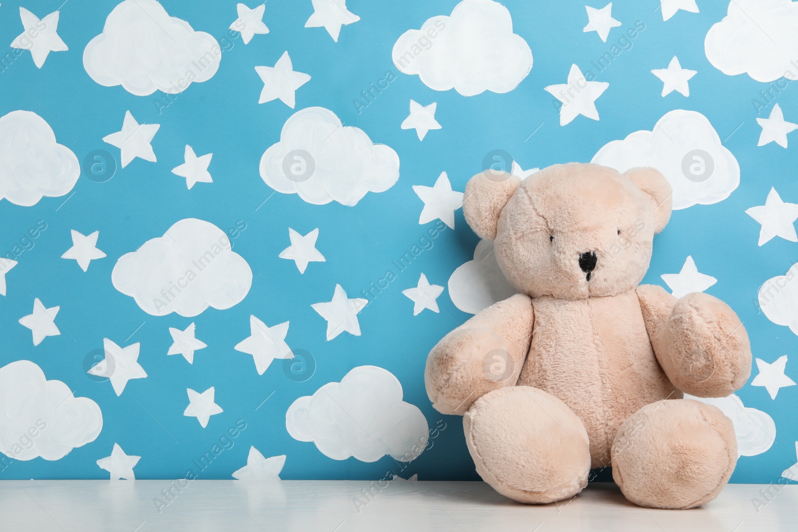 Photo of Teddy bear on white wooden table near wall with painted blue sky, space for text. Baby room interior