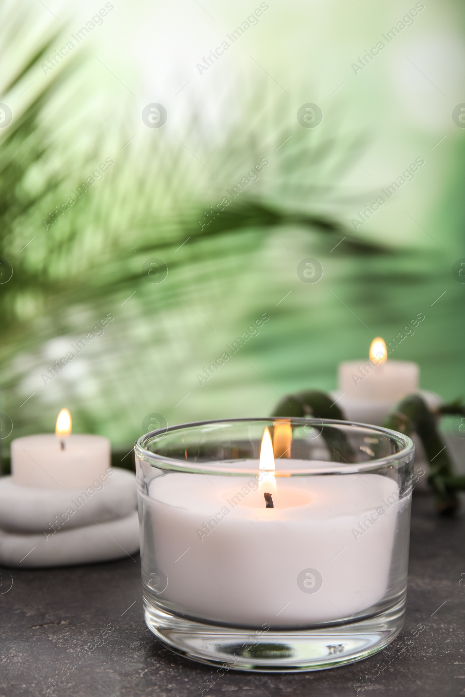 Photo of Burning candle in glass holder on grey table against blurred green background, space for text