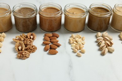 Photo of Tasty nut butters in jars and raw nuts on white marble table, closeup