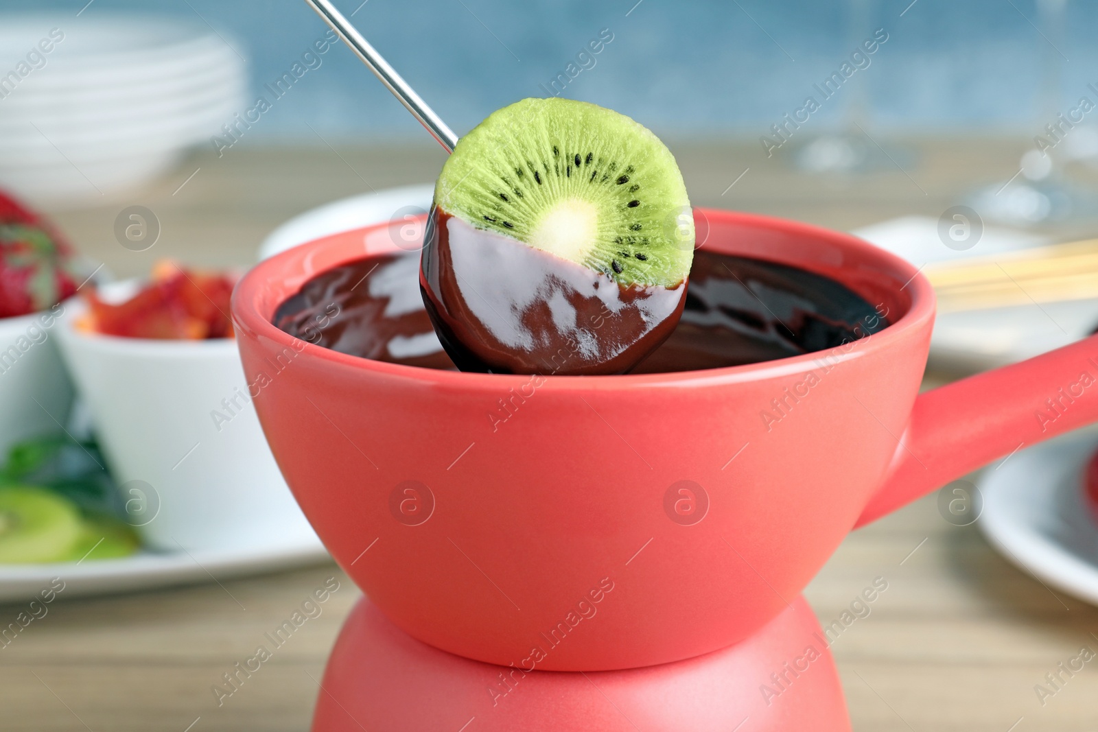 Photo of Dipping kiwi slice into pot with chocolate fondue on table, closeup