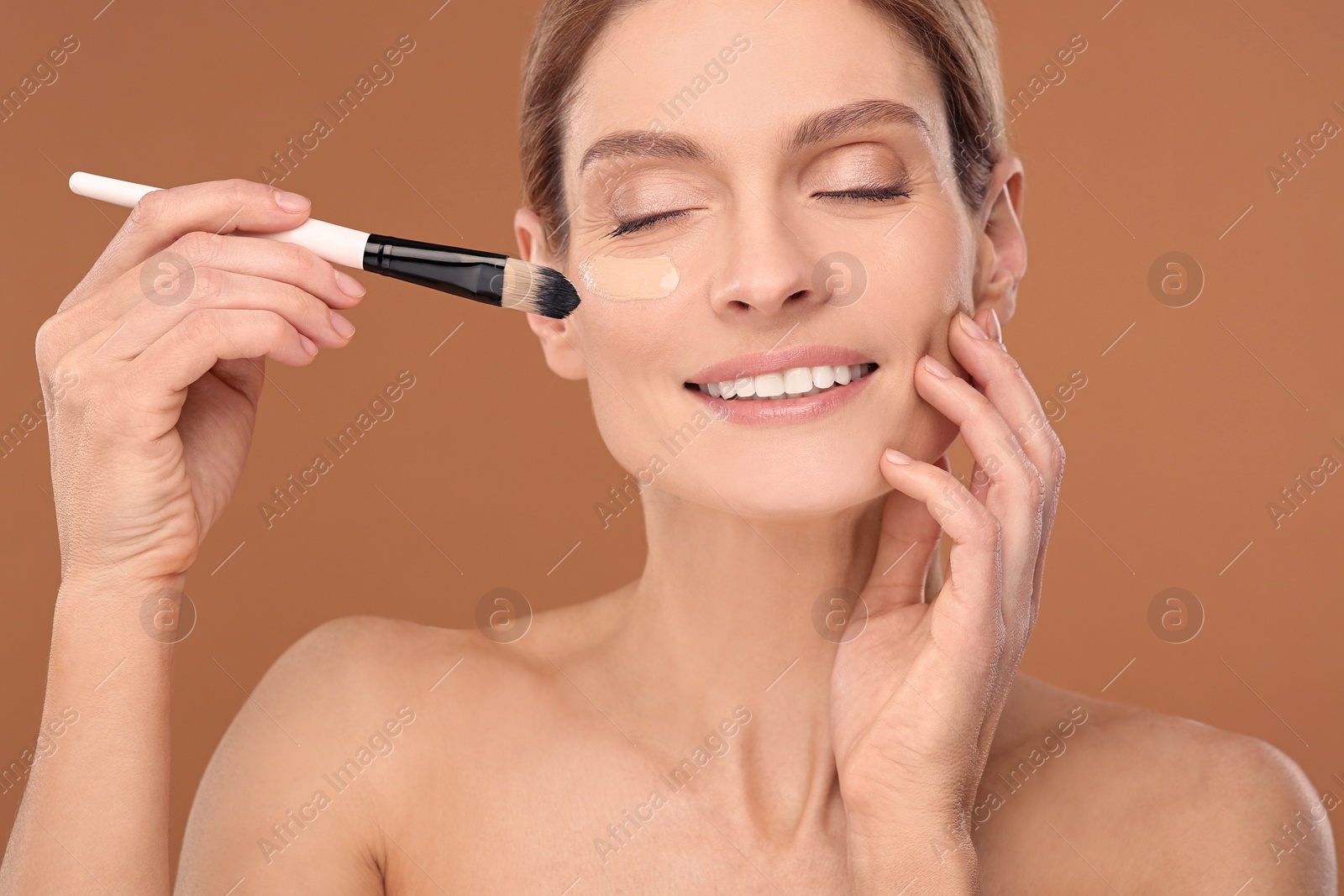 Photo of Woman applying foundation on face with brush against brown background