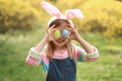 Easter celebration. Little girl in bunny ears covering eyes with painted eggs outdoors